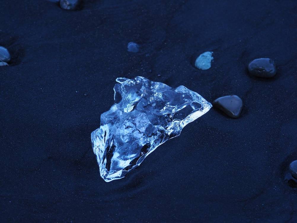 Close-up of a transparent ice chunk resting on the black sand of Diamond Beach in Iceland, showcasing the unique contrast between the ice and the volcanic sand.