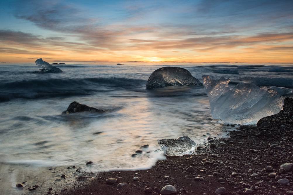 Golden sunrise illuminating the ice chunks scattered along the black sand of Diamond Beach in Iceland, with the waves gently lapping against the shore