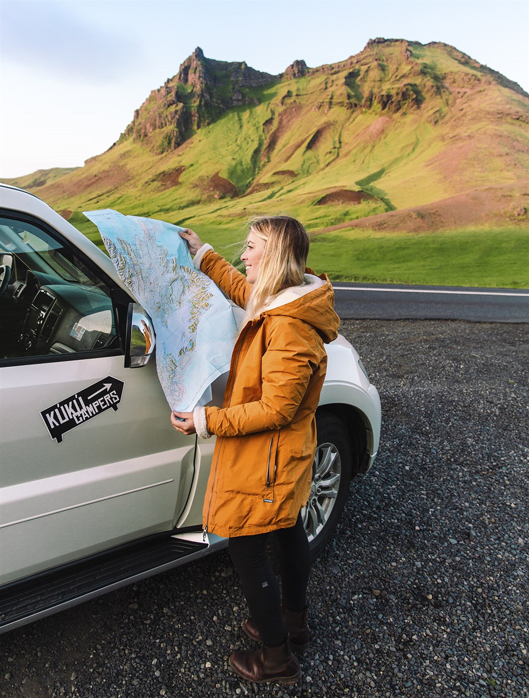 Traveler checking a map next to a KuKu campervan during a scenic Iceland road trip surrounded by green mountains.