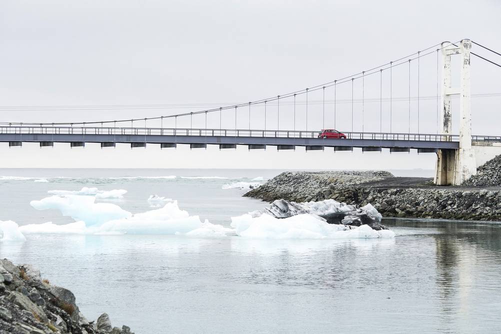 A picturesque view of the bridge near Jökulsárlón Glacier Lagoon in Iceland, with chunks of ice floating towards the open ocean, framed by the serene Icelandic landscape.