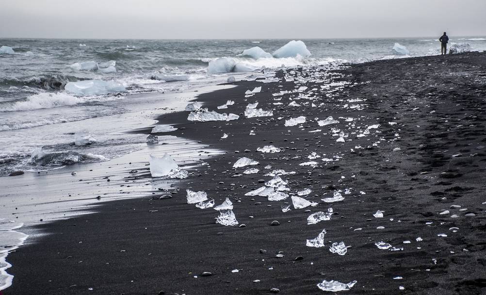 A tranquil scene of small ice fragments scattered along the black sand shoreline of Diamond Beach in Iceland, with waves gently touching the shore.