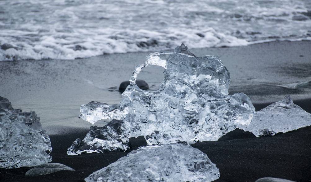 A stunning close-up of a transparent ice chunk resting on the black sand of Diamond Beach in Iceland. The contrast between the crystal-clear ice and the dark volcanic sand creates a mesmerizing visual effect