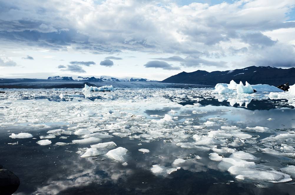 Scenic view of Jökulsárlón Glacier Lagoon in Iceland, featuring floating icebergs and a serene reflection of the cloudy sky on the calm water, framed by distant mountains.