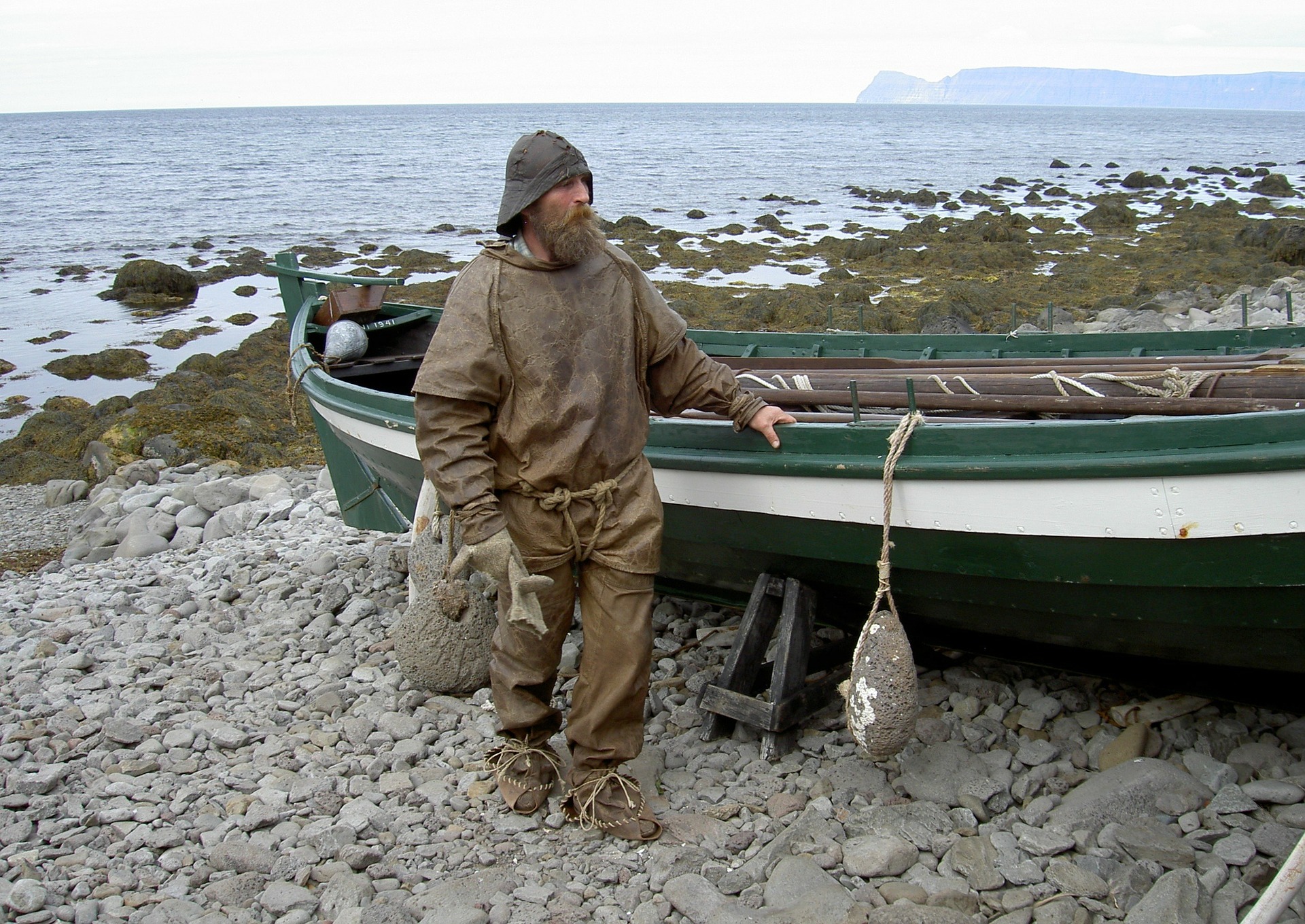 A man dressed in old viking clothes, leather, green wooden boat behing, sea on the background