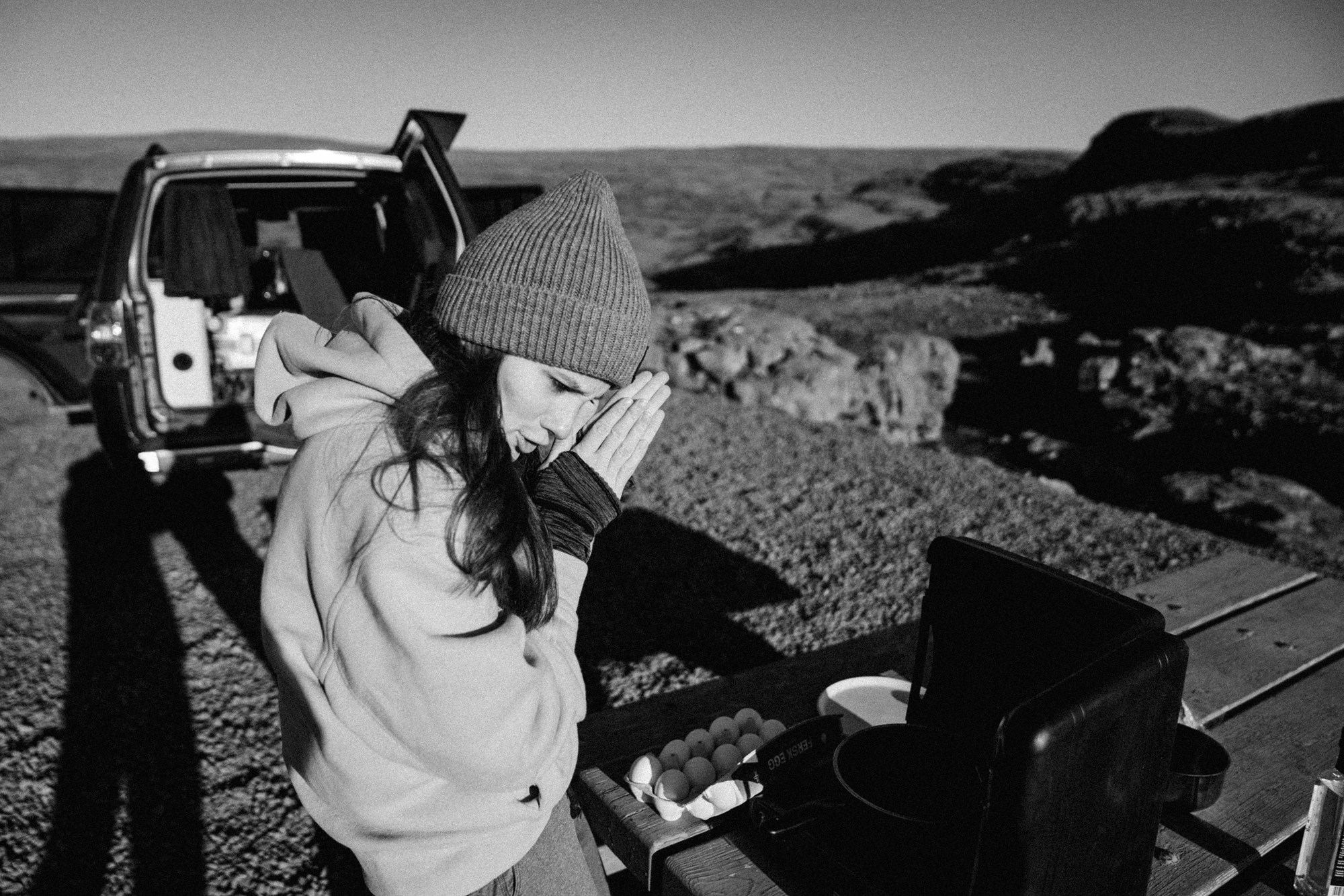 A camper warms their hands before cooking breakfast outdoors, capturing the classic Iceland routine where the stove heats the food and the traveler heats themselves.