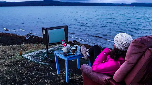A traveler lounges by an Icelandic shoreline with a vintage TV and a cup of something warm, proving that even the middle of nowhere can feel like prime time.