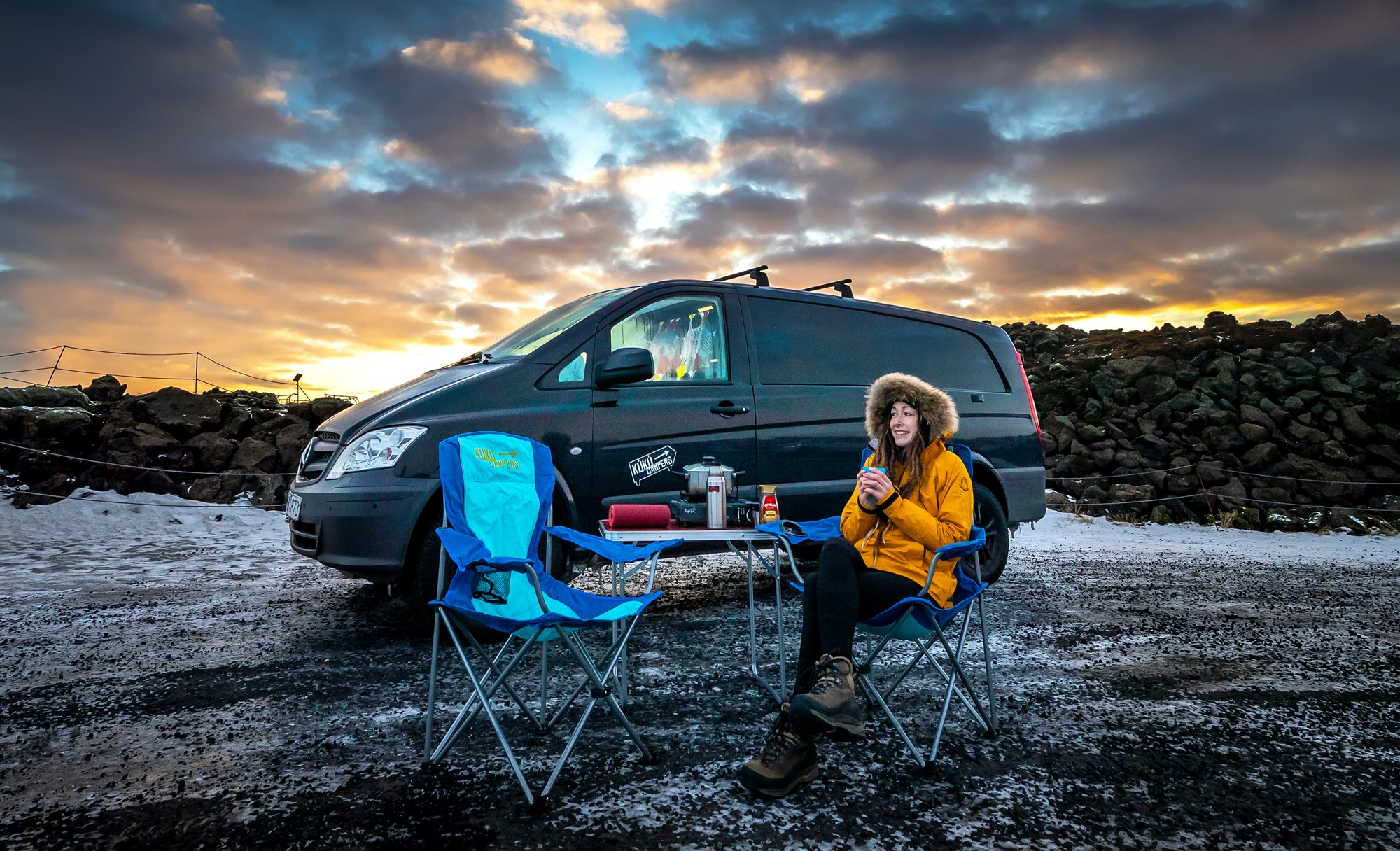 Woman sitting in front of a camping chair eating breakfast on a camping table in front of a black Mercedes Vito van.